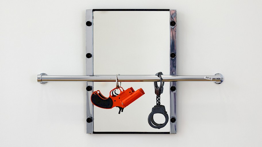 A mirror with a silver frame is attached to a white wall. In front of it, just below the center, is a metal bar, which is also attached to the wall. A small orange revolver and black handcuffs hang from it in front of the mirror.  Image: Cady Noland, The Mirror Device, 1987, photo: Axel Schneider.