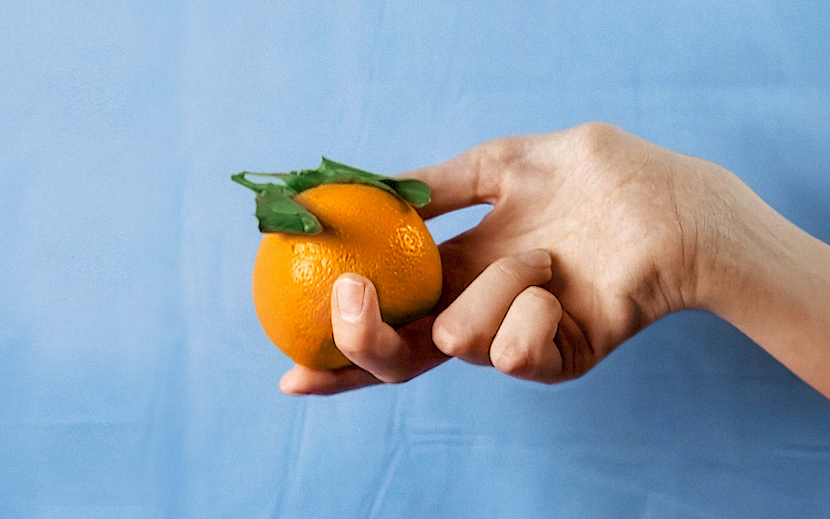 Michelle Miles, hand model, 2018, Filmstill. Bildbeschreibung: Eine Frauenhand hält eine leuchtende Orange vor einem hellblauen, leicht zerknitterten Hintergrund aus Stoff.