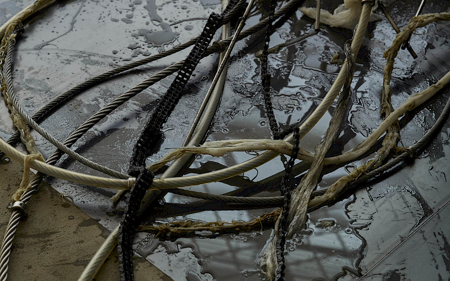 On a light gray concrete floor lies a steel plate on which a liquid has formed puddles. Dirty cords, chains and hoses lie interwoven in them. Image: Mire Lee, Horizontal Forms, 2020 (detail), photo: Yonje Kim