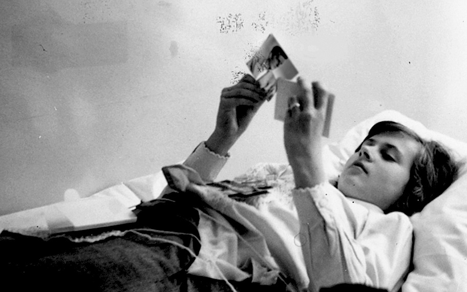Image description: You can see a black and white photograph of a young person lying in a bed. She is looking at two photographs with her head on the pillow. The rest of the photographs are on her upper body. The background is bright and in the foreground is the shadowy image of the edge of the bed. Credit: Rosemarie Trockel, First Chlamydia, 1966/2017, Private Collection, © The artist & VG Bild-Kunst, Bonn 2022