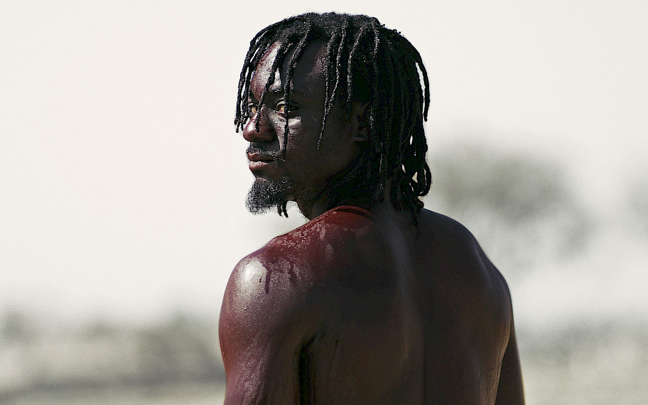Image descritpion: You can see a middle-aged male person who has turned his naked and blood-covered back to the person looking at him. The person looks at us over his left shoulder, so that his profile becomes visible. He wears a black beard and chin-length black hair. A few trees are silhouetted in the background.
credit: The Critics Company, One Can Only Hope and Wonder, 2023, film still