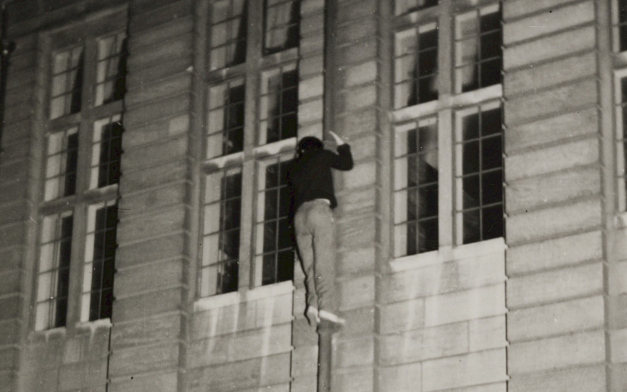Image description: One can see a black and white photo of a building facade. In the middle of the picture a person is holding on to a pipe of the facade. Credit: The Night Climbers of Cambridge, Untitled, 1930s (detail), MUSEUM MMK FÜR MODERNE KUNST, © Thomas Mailaender, photo: Axel Schneider